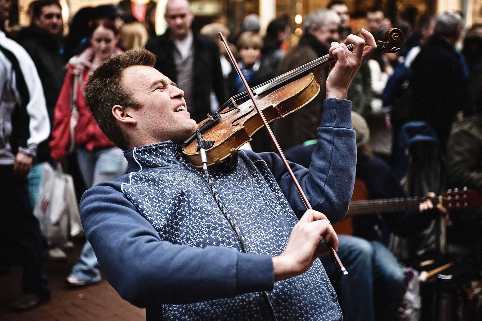 Man playing violin enthusiastically in a crowded street; blue sweater, instrument held up.