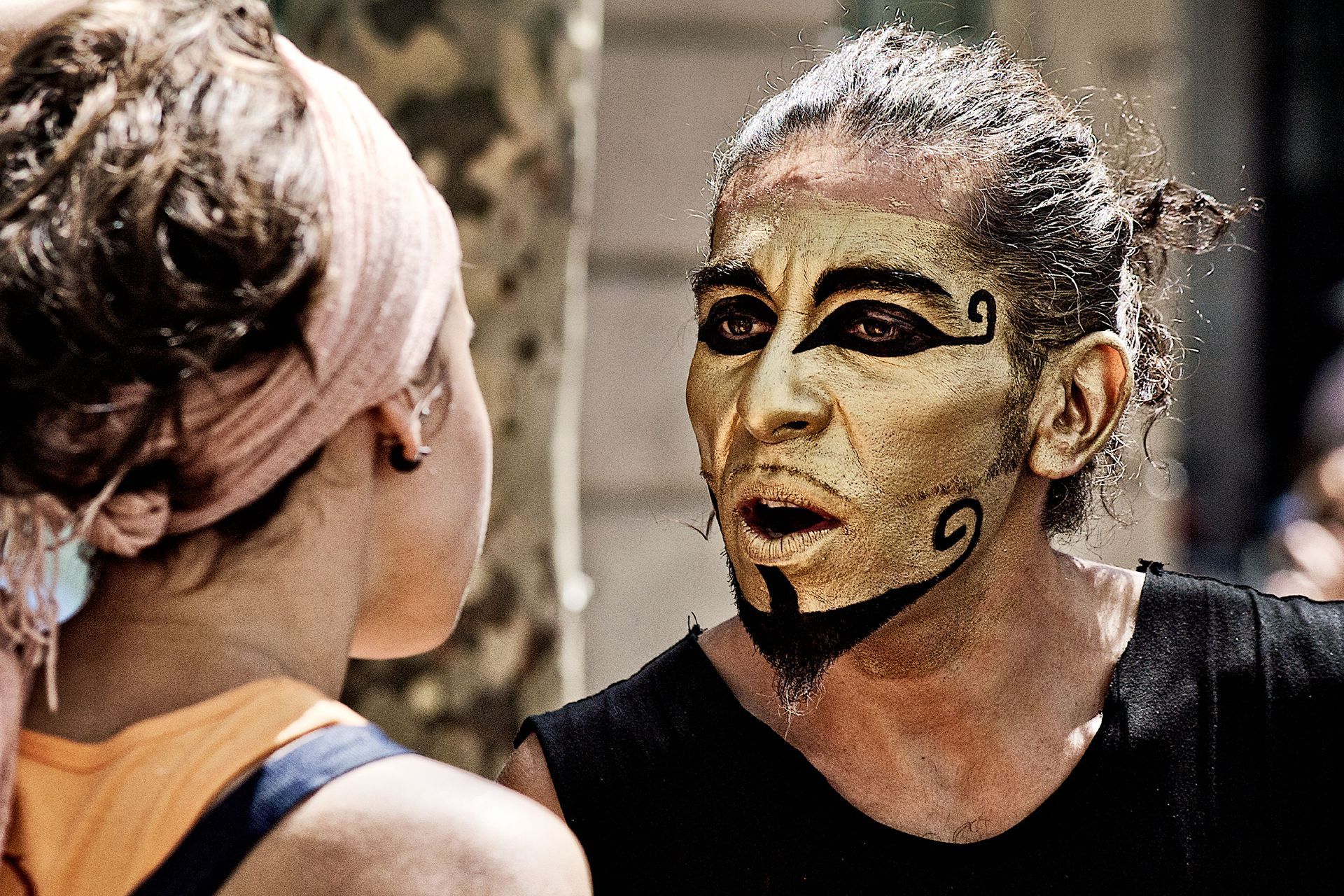 Man with gold face paint and black lines, talking to woman with headscarf.