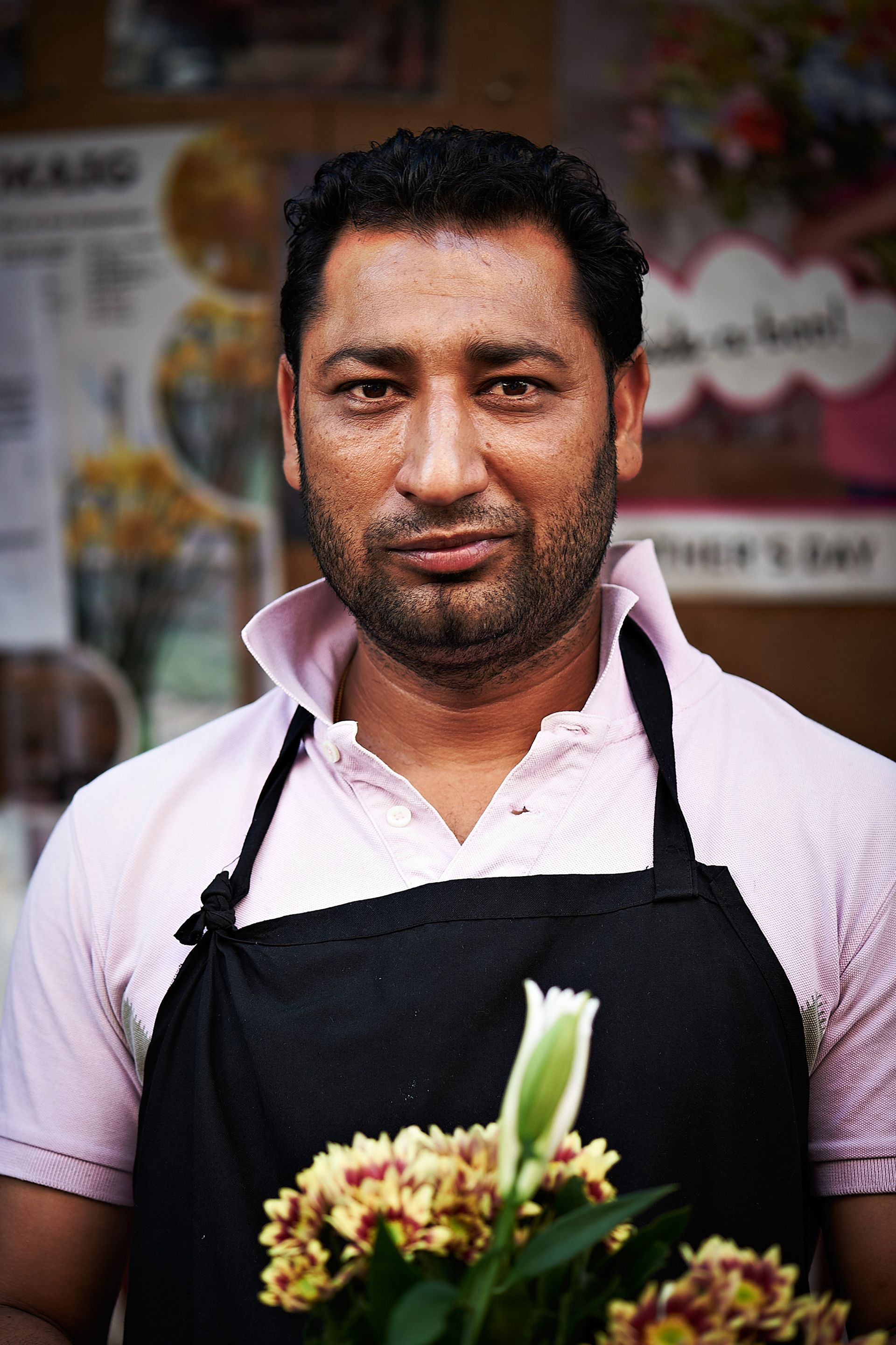 Man in pink shirt and black apron holding flowers in front of a flower shop.
