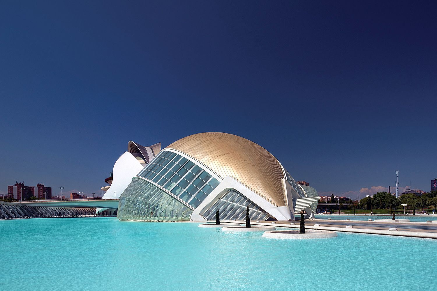Modern building with a golden dome and glass facade reflected in turquoise water against a blue sky.