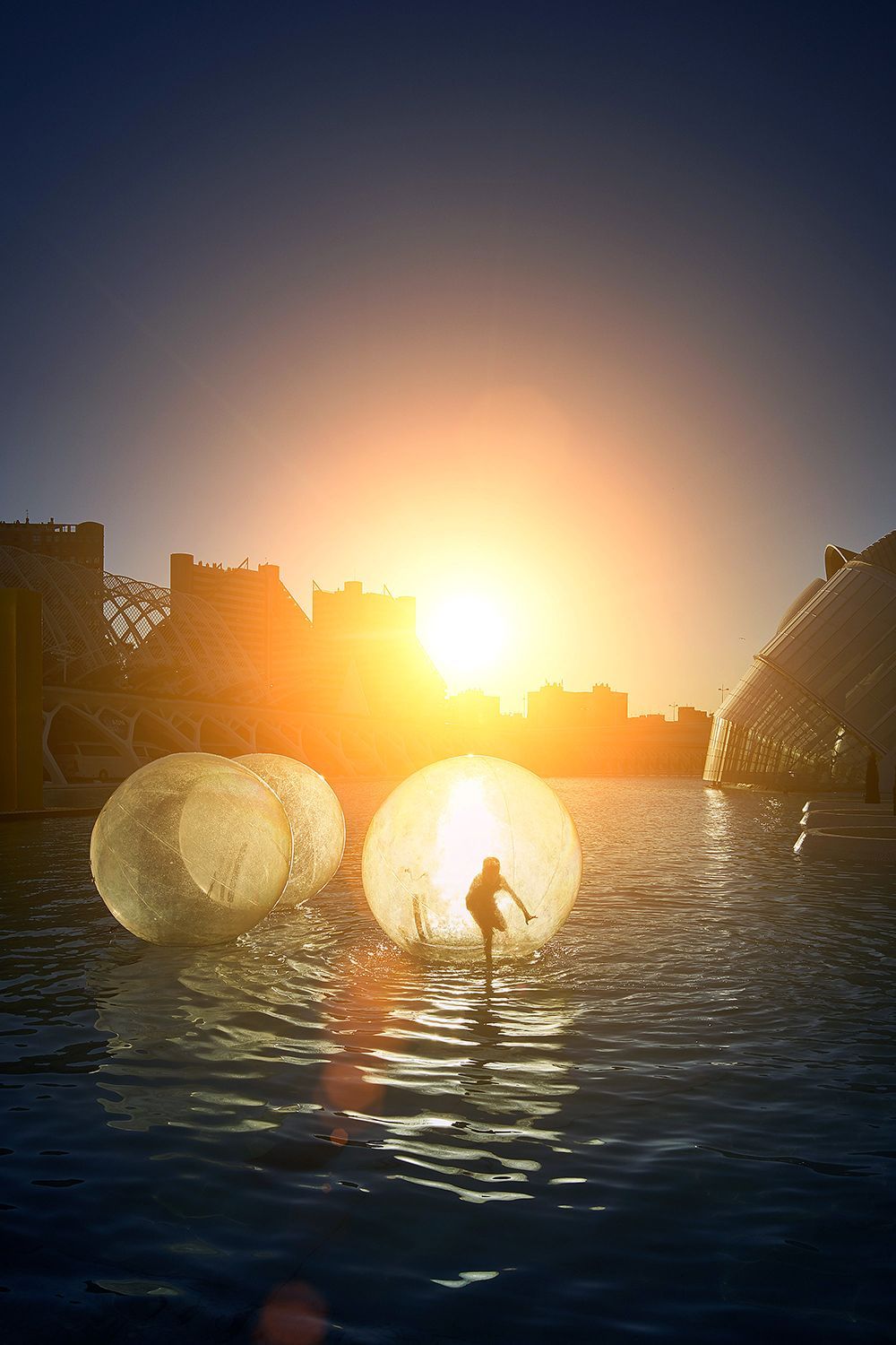 Person in water, standing inside an inflatable ball at sunset, with two similar balls. City skyline in background.