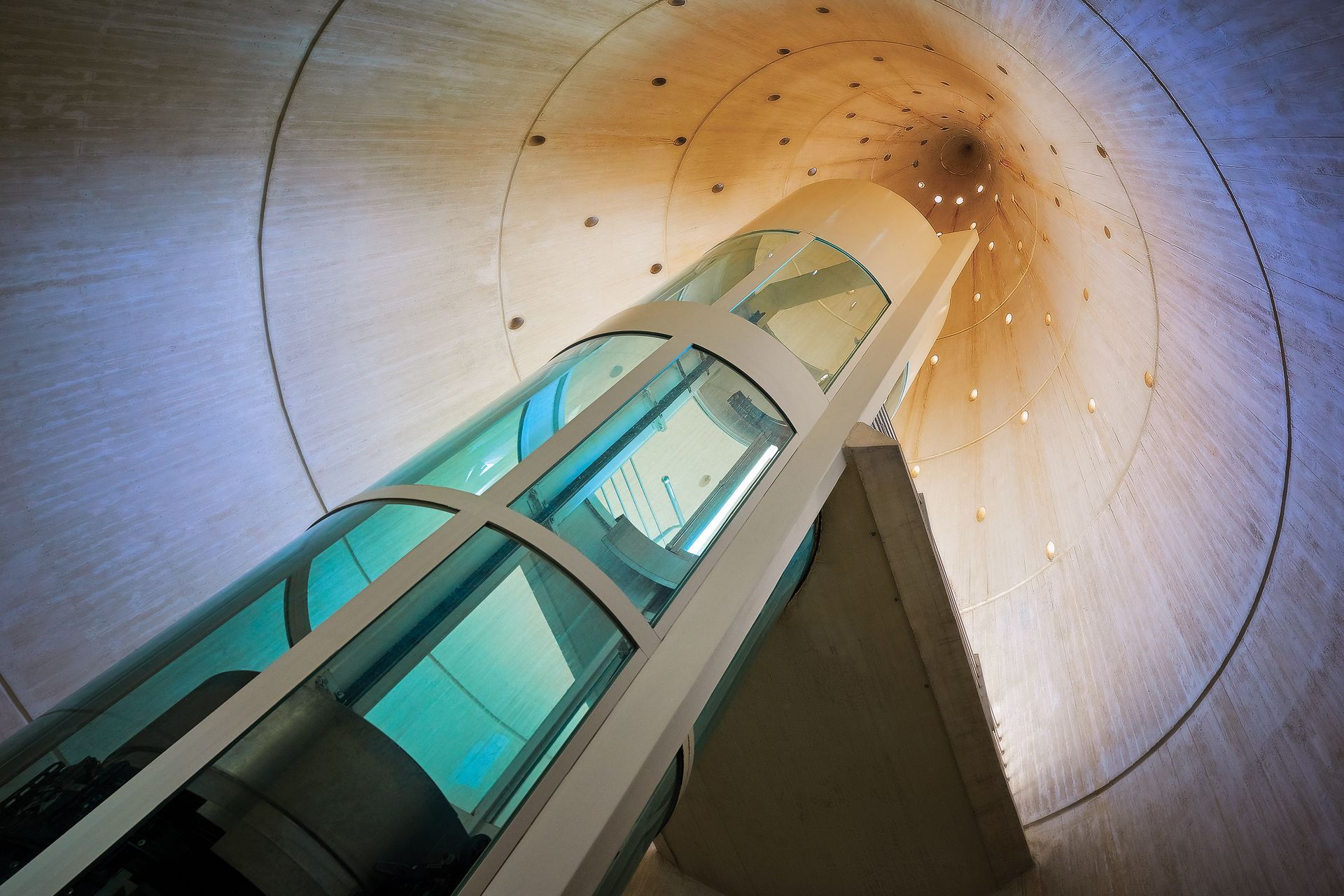 Glass elevator ascends a concrete circular shaft, viewed from below.