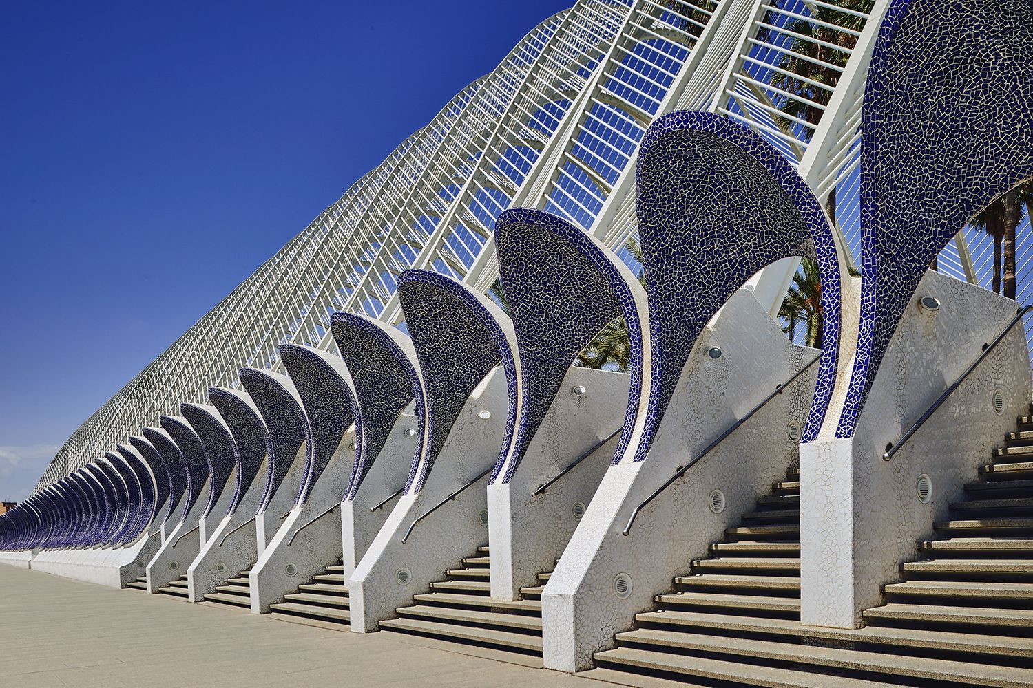 Modern building with white angled supports, blue mosaic details, and a glass roof against a blue sky.