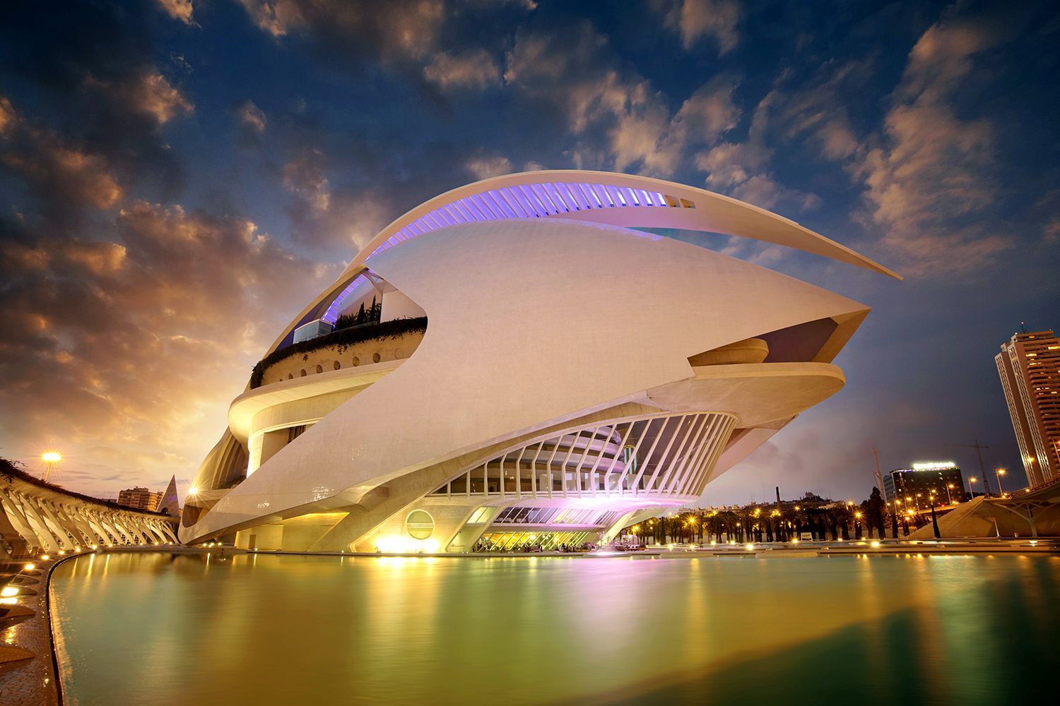 Futuristic white building with curved shapes, lit from below, reflected in water under a dramatic sunset.
