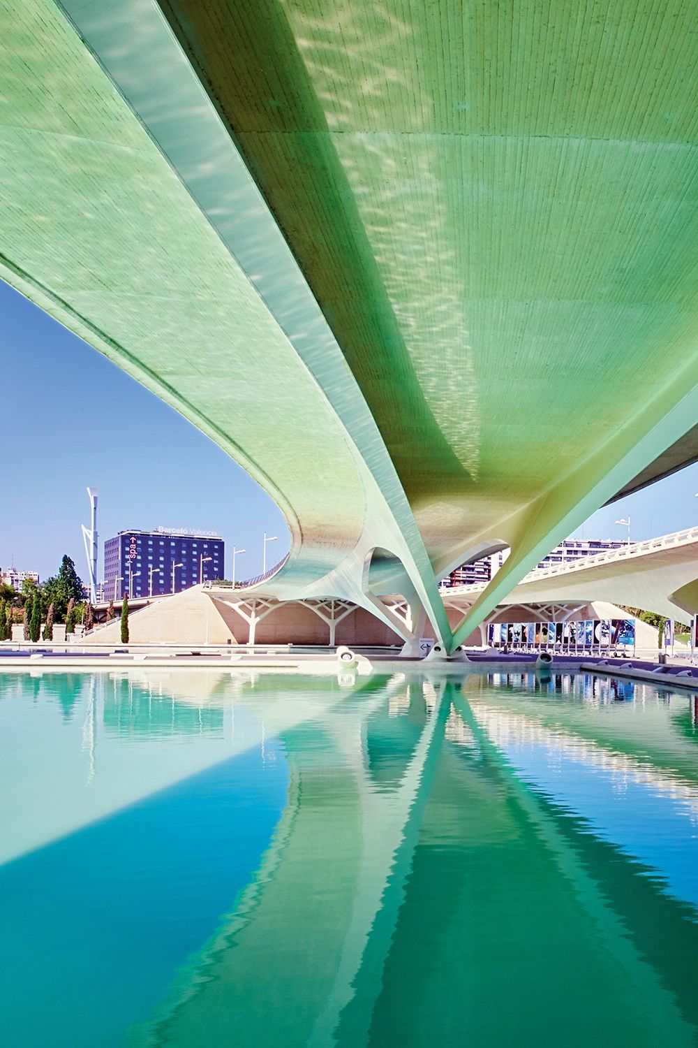 Modern arched bridge over teal water, reflecting the structure and a sunny sky.