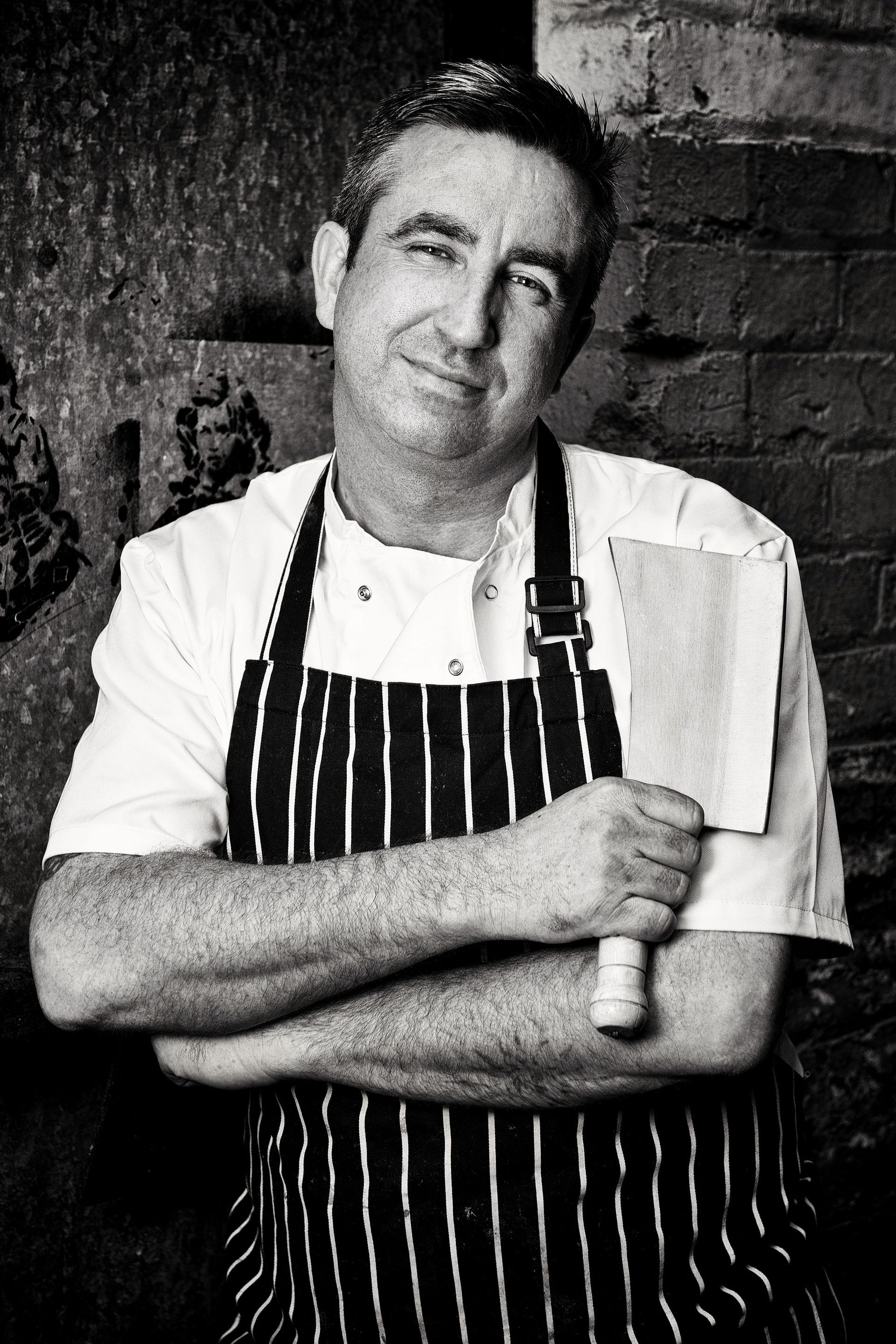 Chef in striped apron holding a cleaver, smiling, standing against a brick wall.