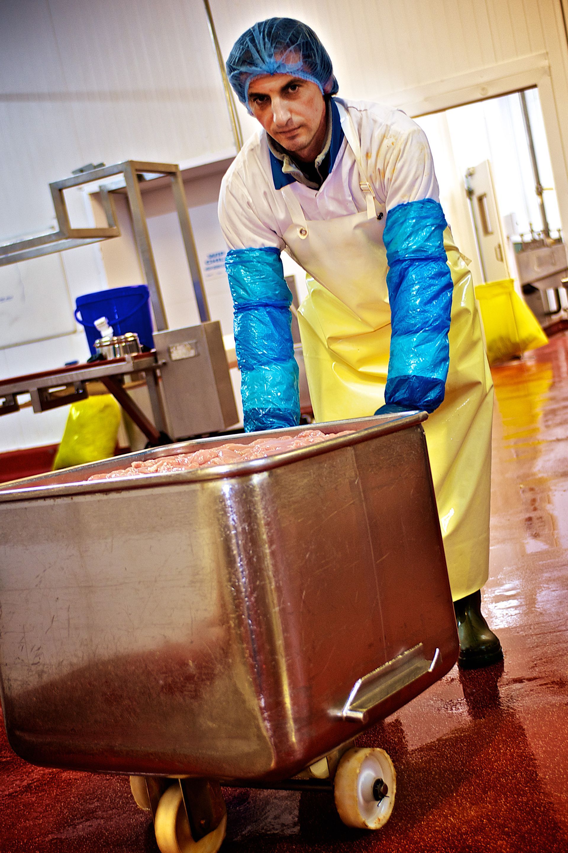 Man in apron and protective sleeves pushes a large bin on wheels across a red-floored industrial room.