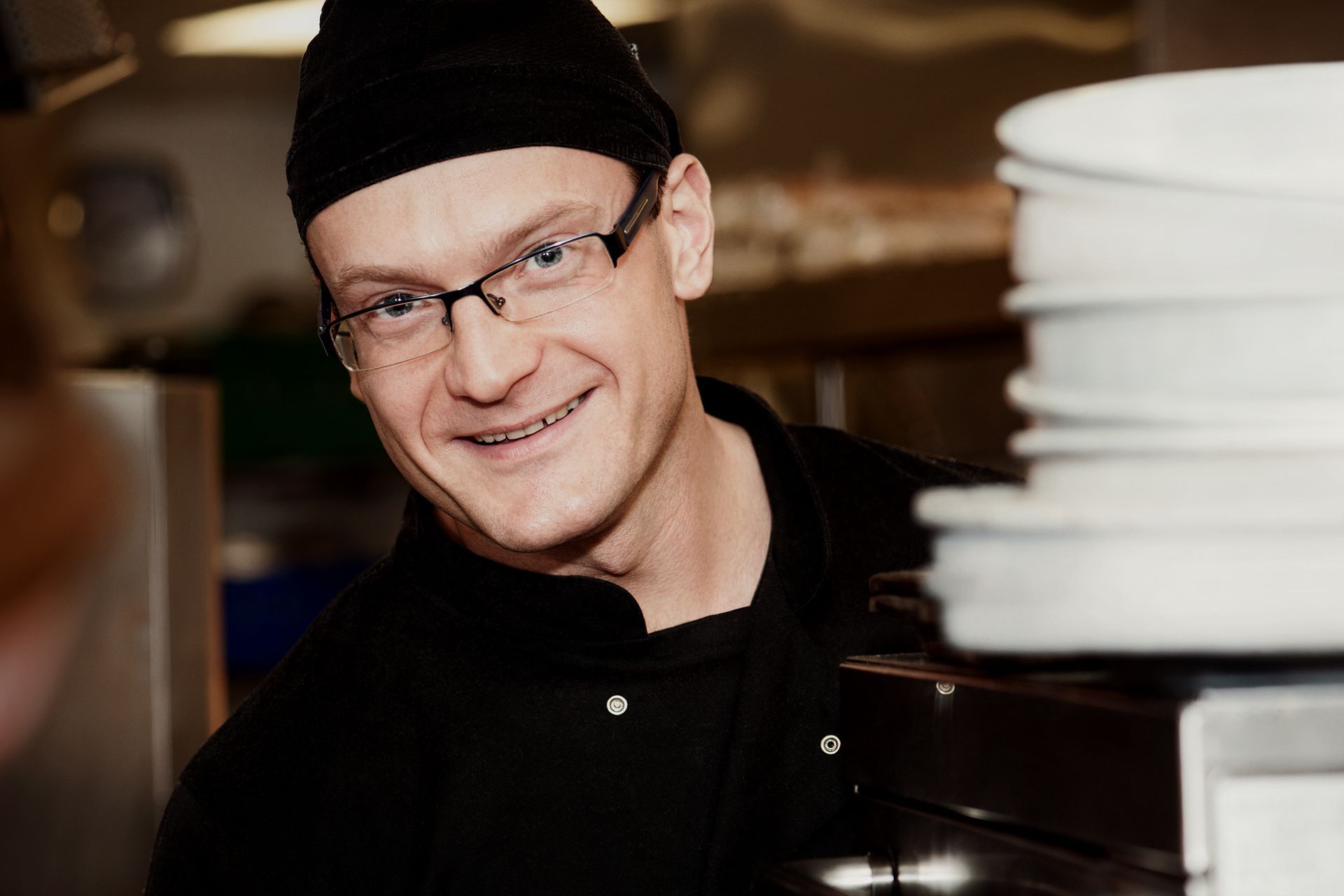 Chef wearing glasses and black hat smiles in a kitchen, holding a stack of plates.