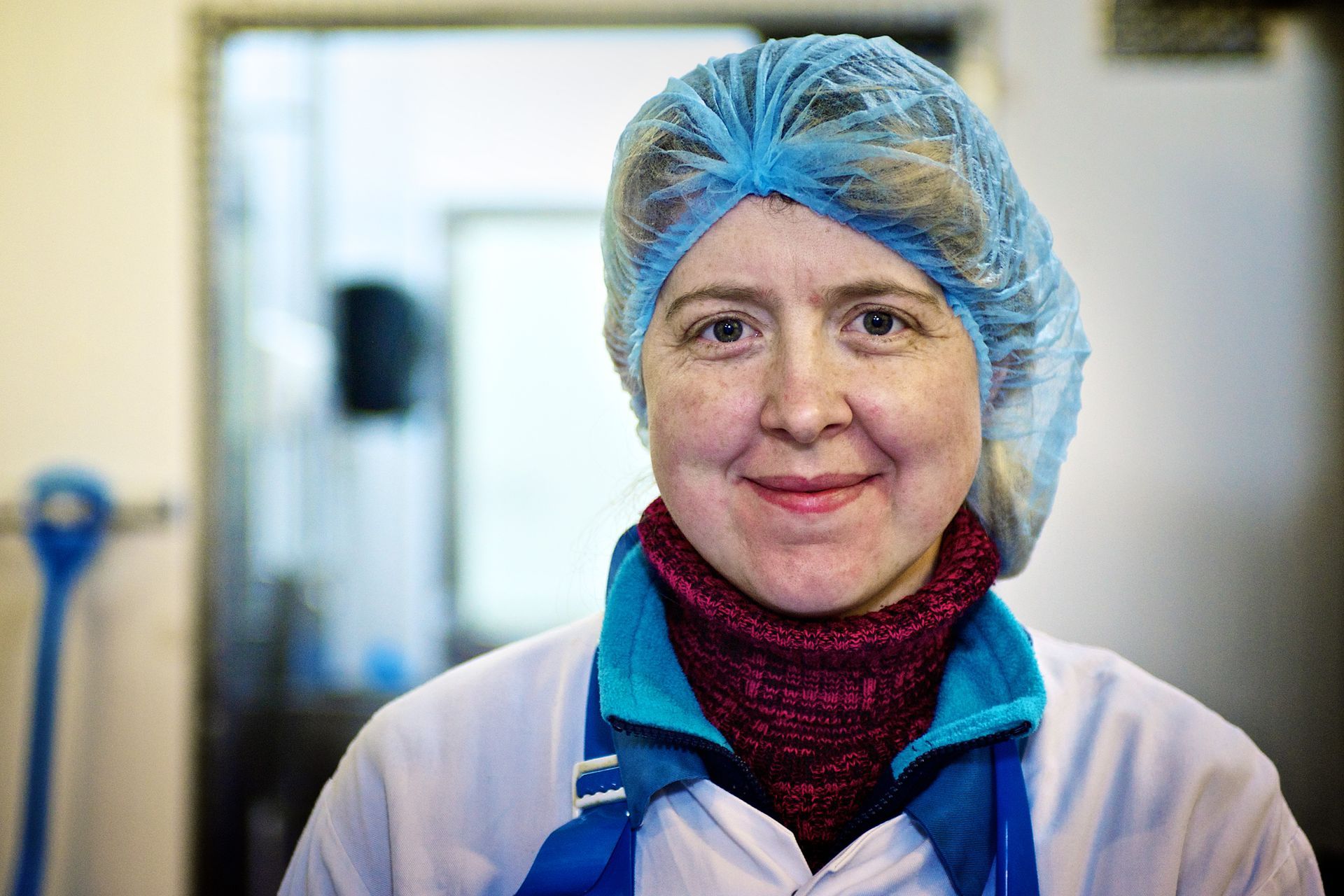 Woman in blue hairnet and white coat smiles, blurry background of a food processing area.