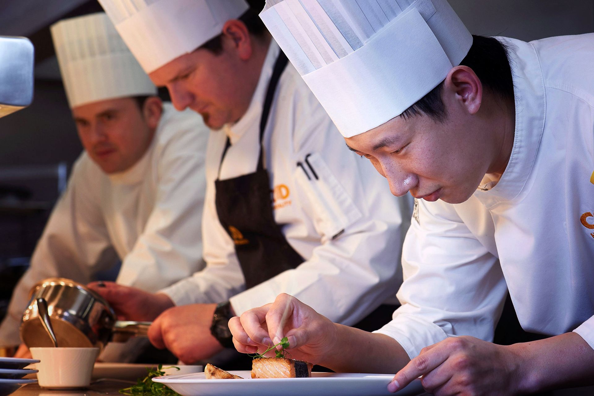 Three chefs in white coats and hats work in a brightly lit kitchen. One plates food, adding garnish.