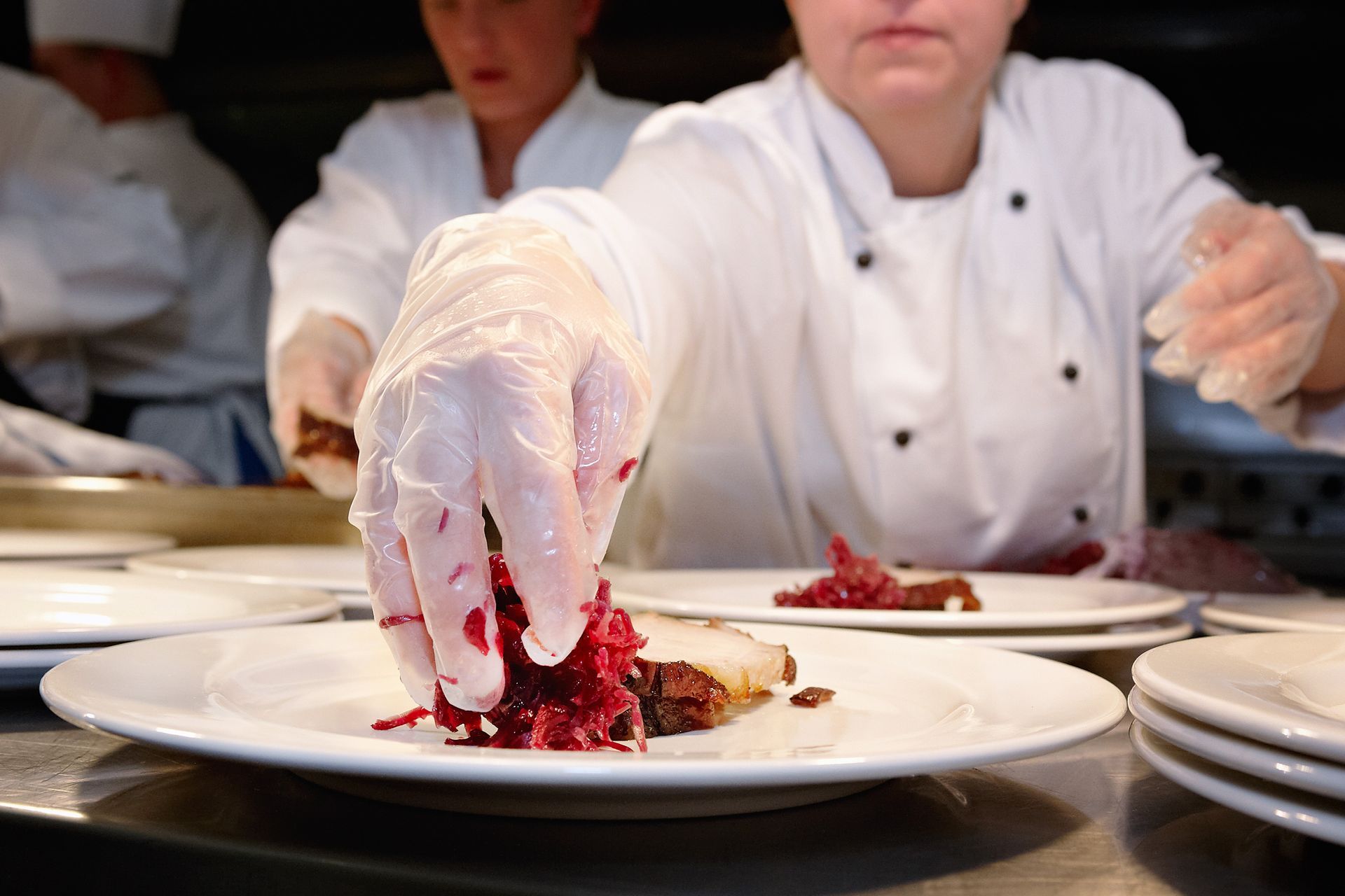Chefs plating food in a busy kitchen; chef with gloved hands placing red slaw on a plate.