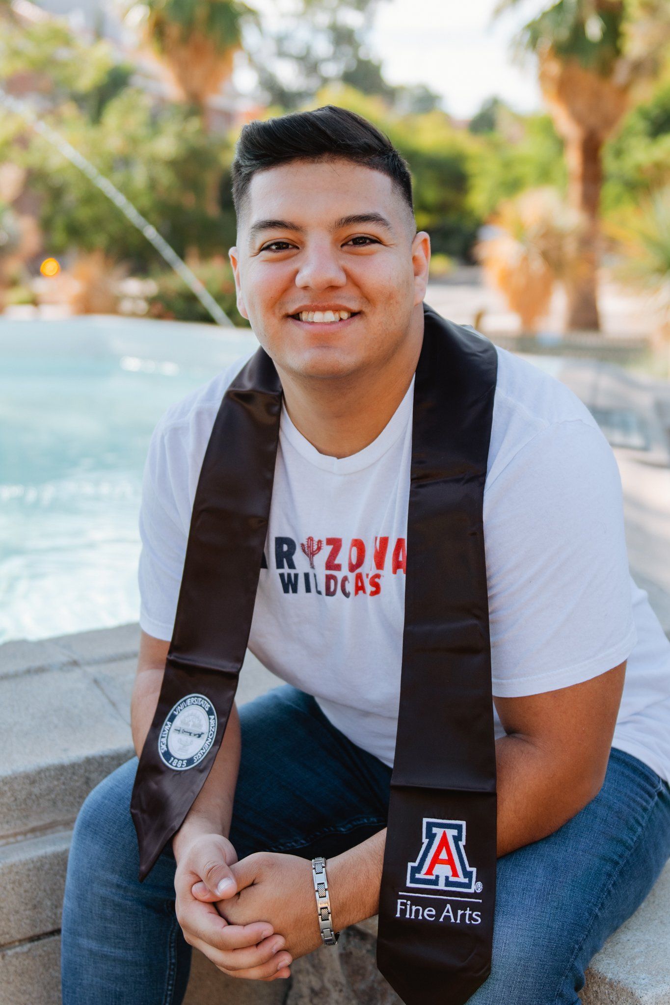 A man wearing a graduation sash is sitting on a ledge next to a fountain.