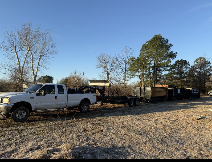 Two black dumpsters are parked in a dirt lot in front of a pile of trash.