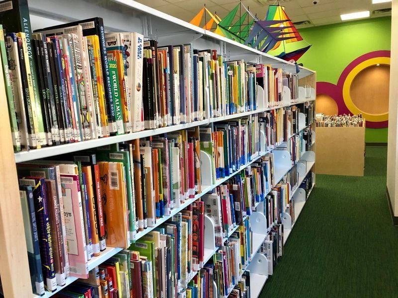 Books on white shelves in a library, green carpet, colorful wall decor, and kites on top shelf.