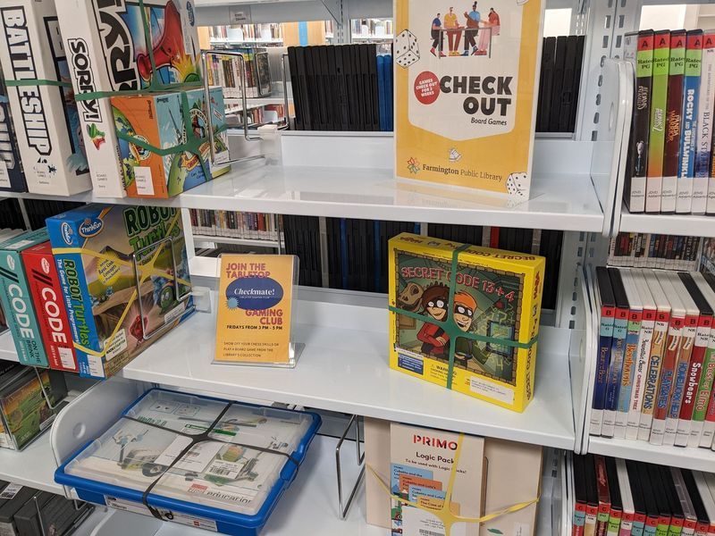Books and board games on library shelves; some are bundled together.