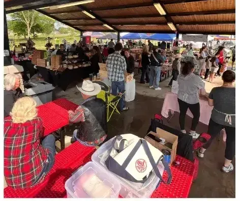 Outdoor market under a covered structure with vendors, shoppers, and people at red picnic tables.