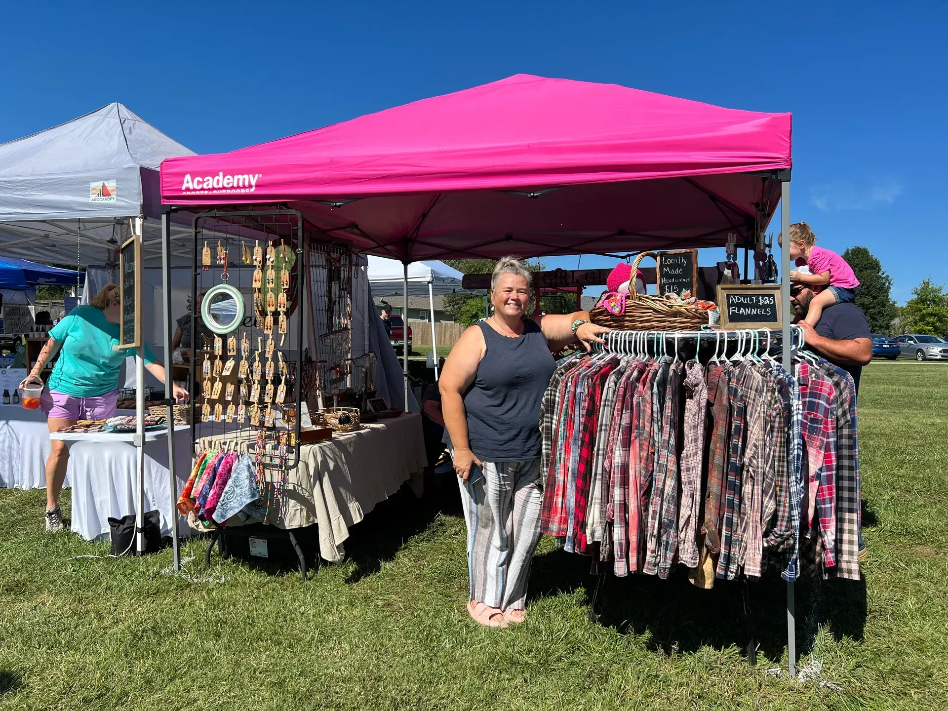 Woman in front of pink tent, at market with clothing and jewelry. A man holds a child on his shoulders. Sunny day.