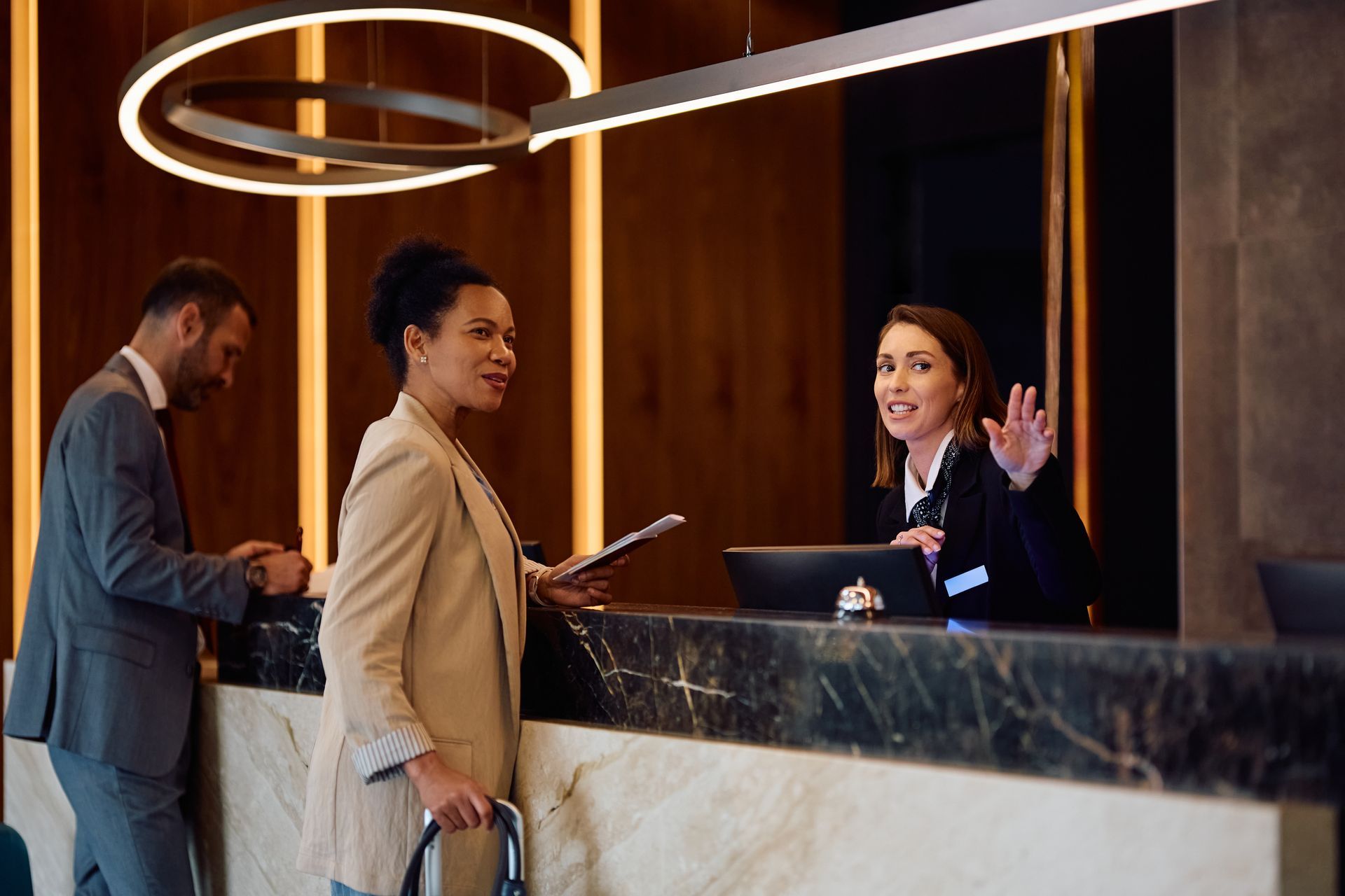 Hotel front desk: Three people; guest, clerk, and other guest. Clerk waves.