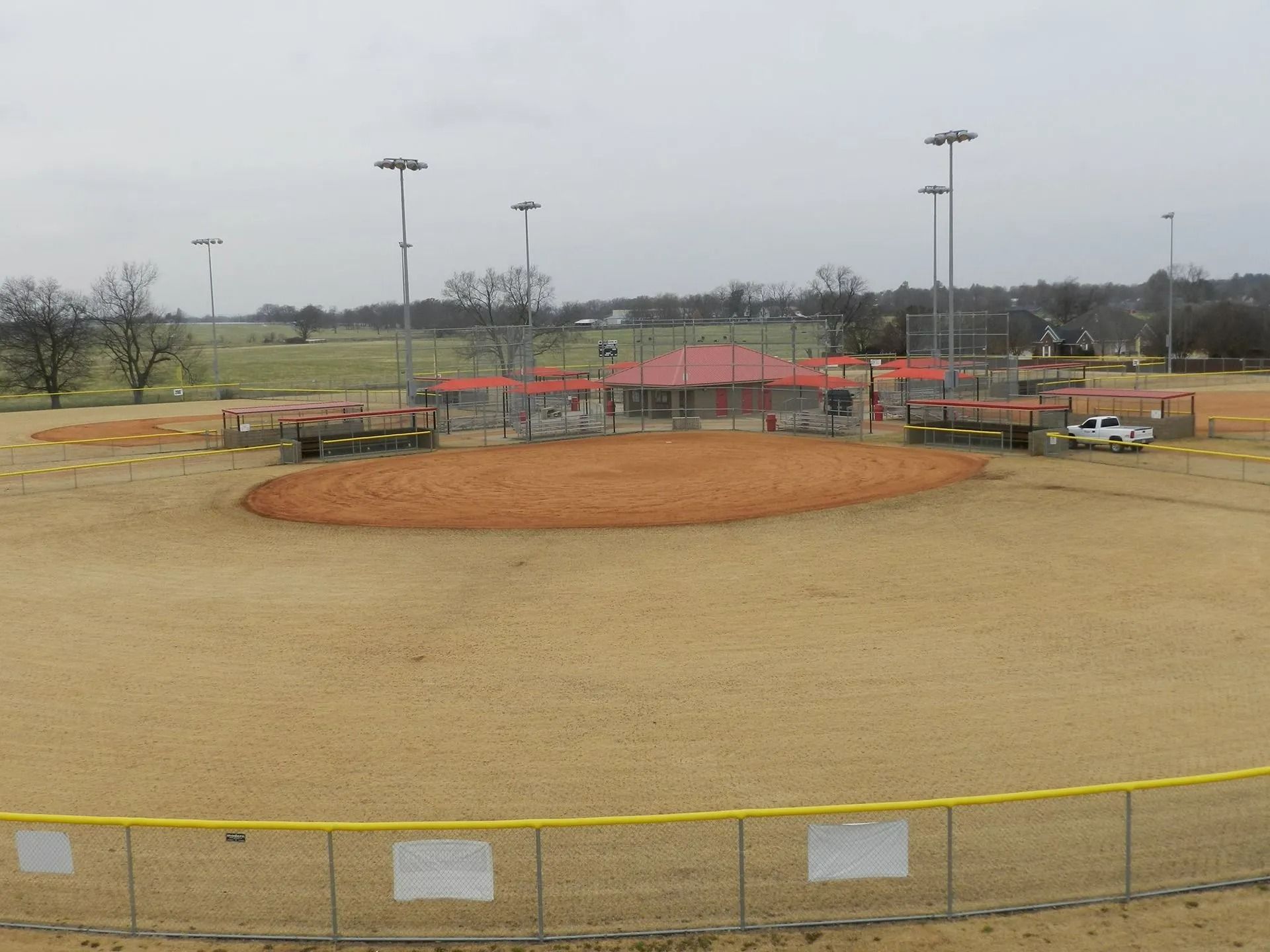 Baseball field on a cloudy day; brown infield, tan outfield, red shelter, yellow fence.