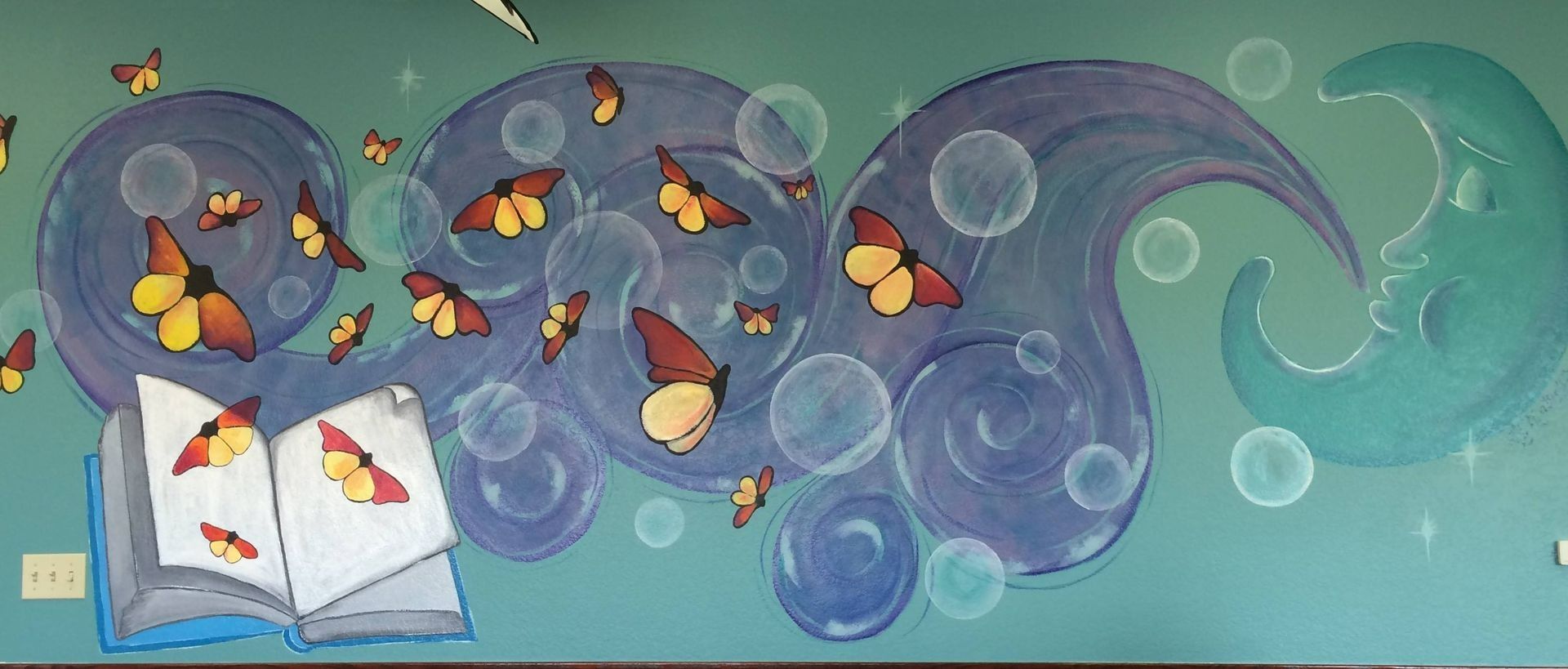 Mural of butterflies emerging from a book, swirling around with bubbles and a moon, painted on a teal wall.
