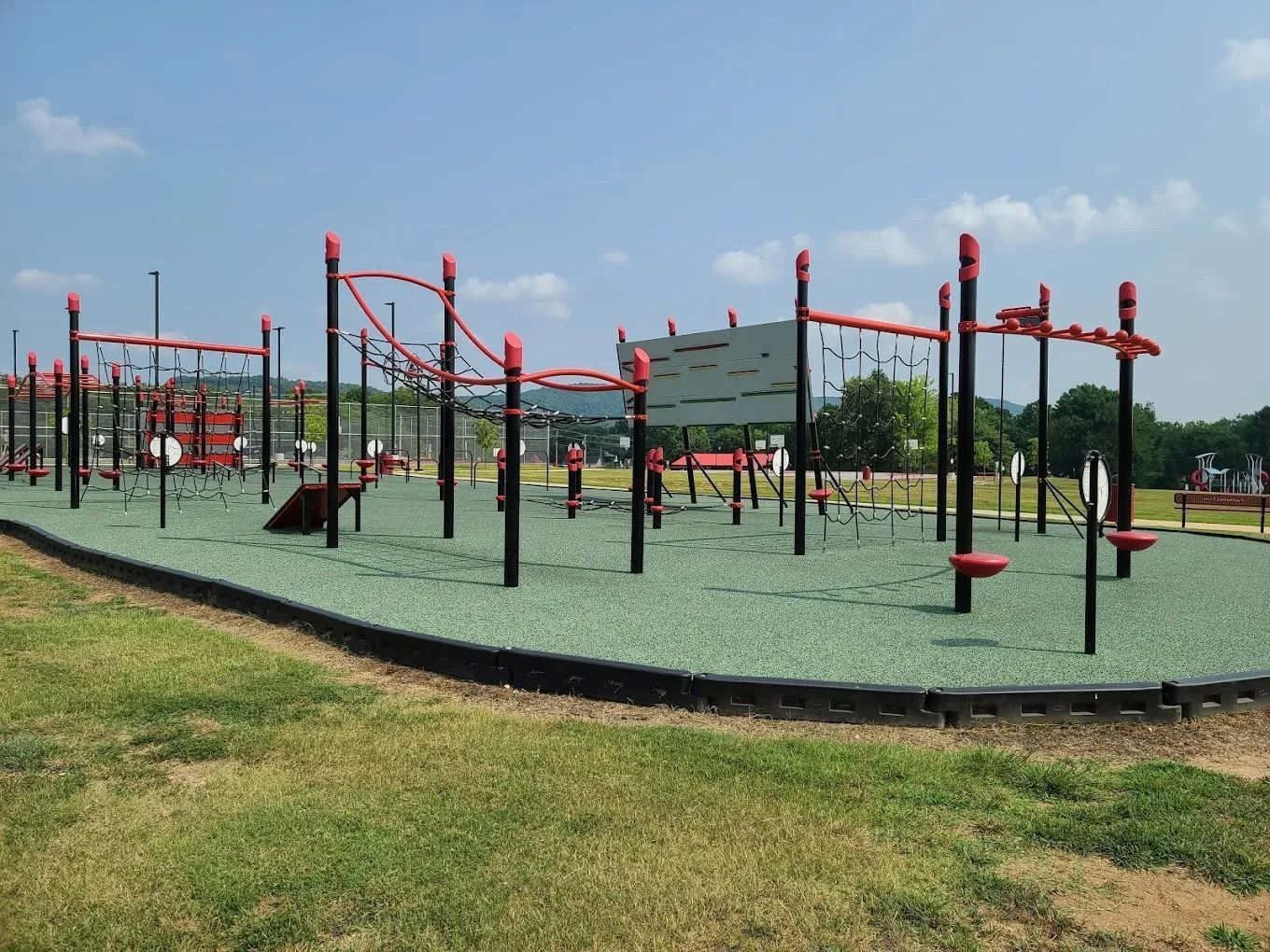 Outdoor playground with black poles, red rails, and green safety surface, under a blue sky.