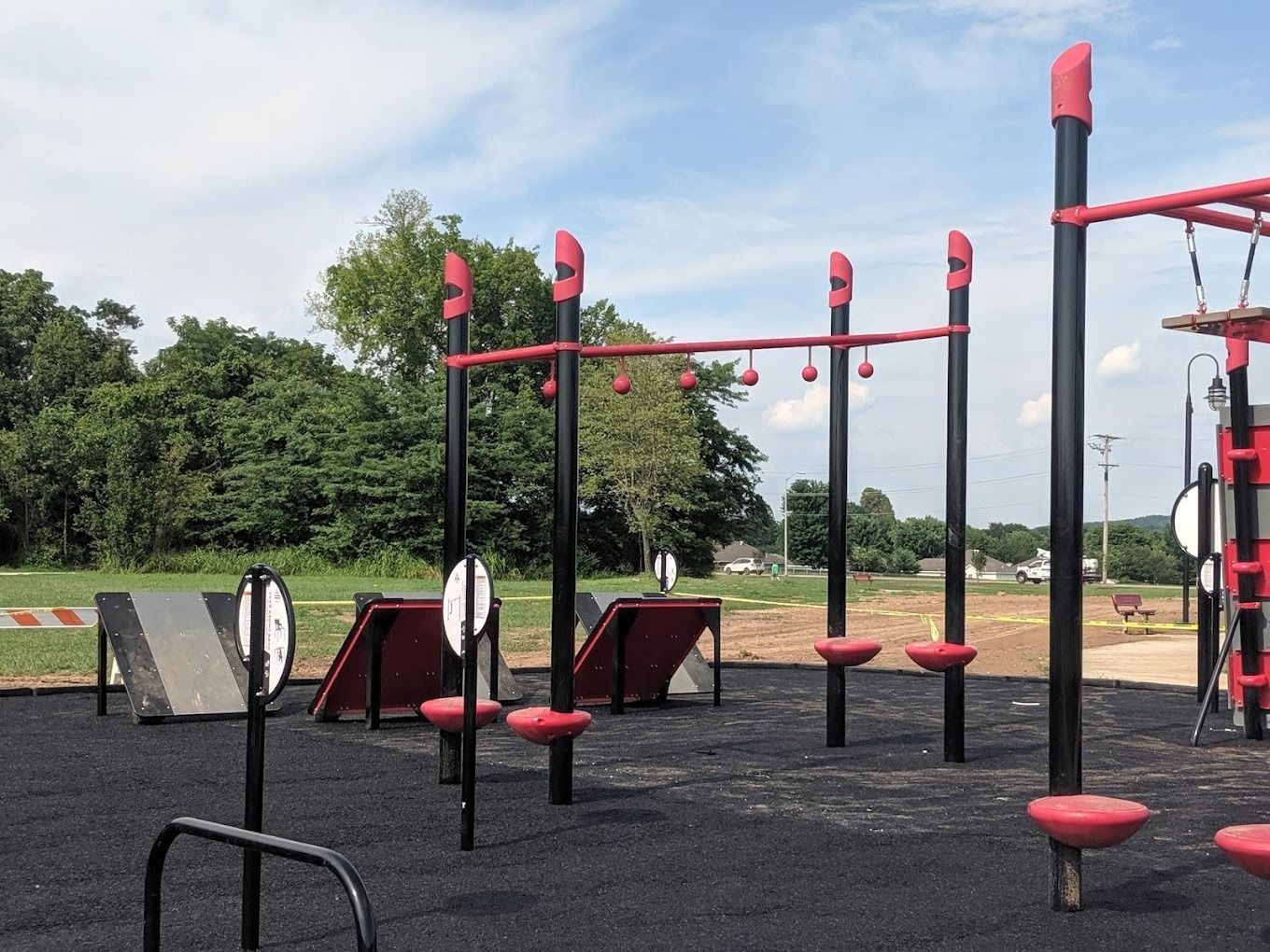 Outdoor fitness park with black and red equipment on a black surface, set against a green tree backdrop and cloudy sky.