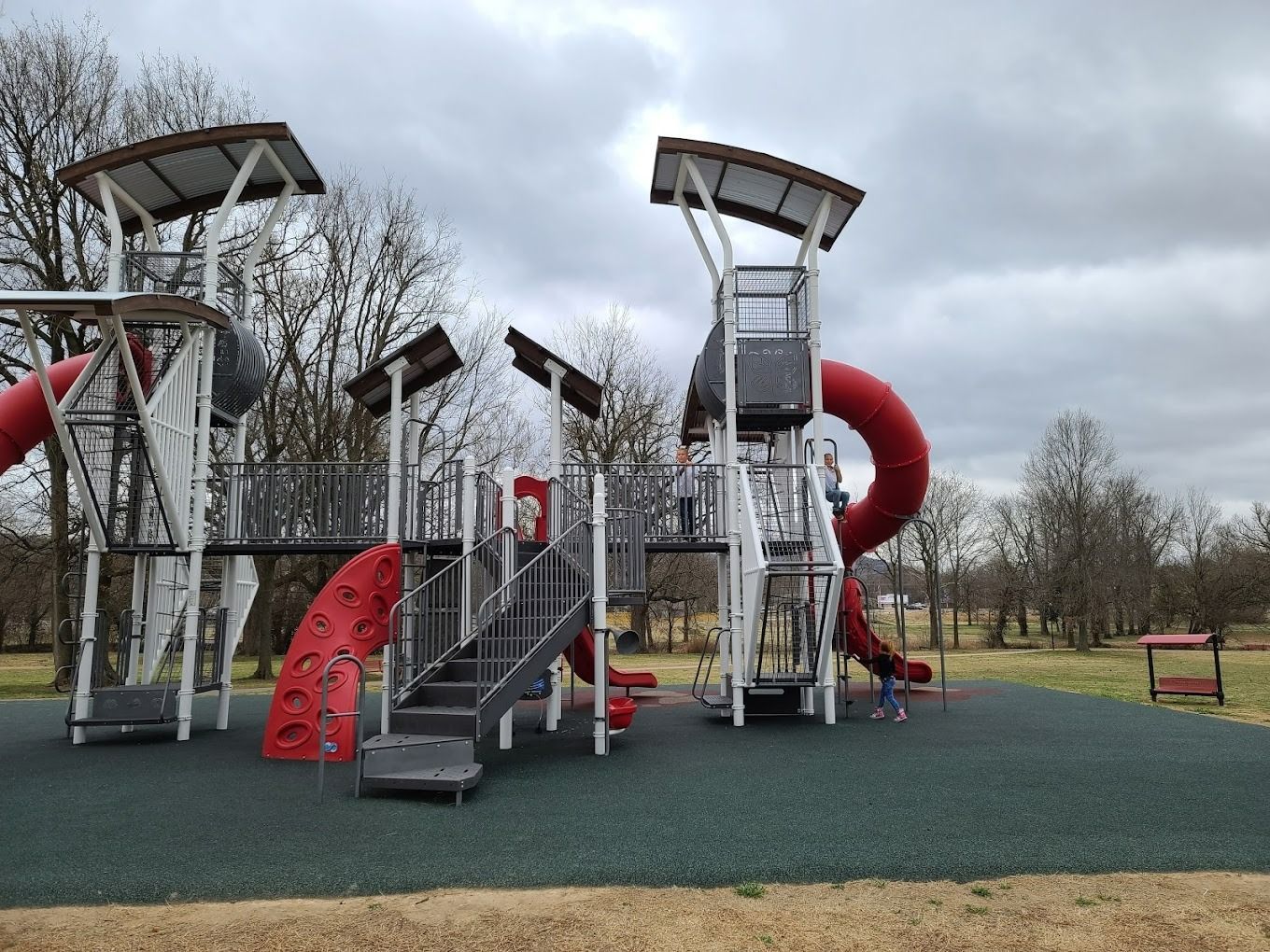 Playground with red slides, climbing structures, and gray metal stairs on a green surface, under a cloudy sky.