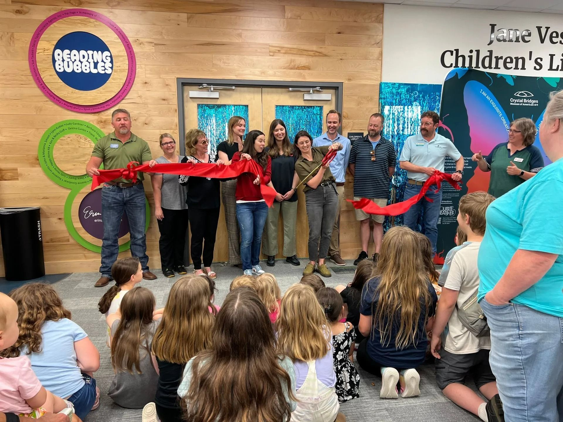 People cutting a ribbon at a library opening, with children watching. The room is brightly lit.