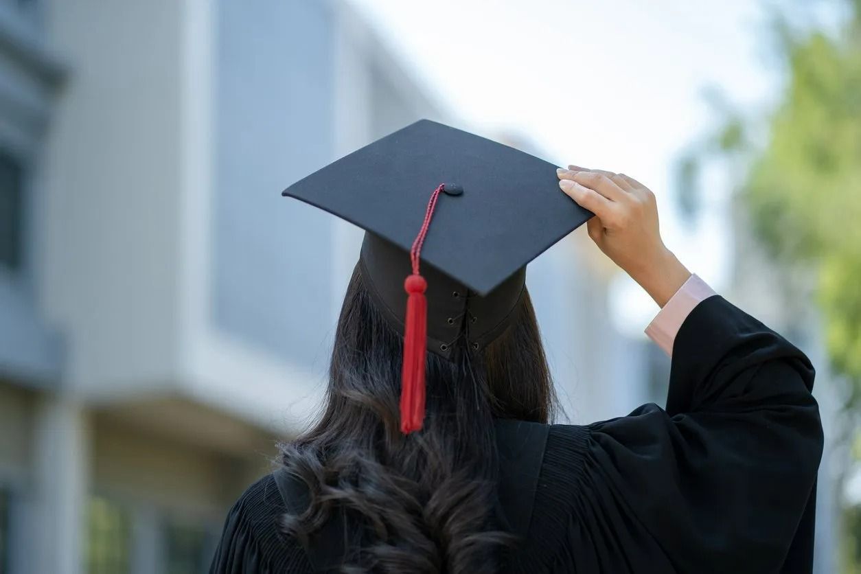 Person in graduation gown, holding mortarboard, looking toward a building.
