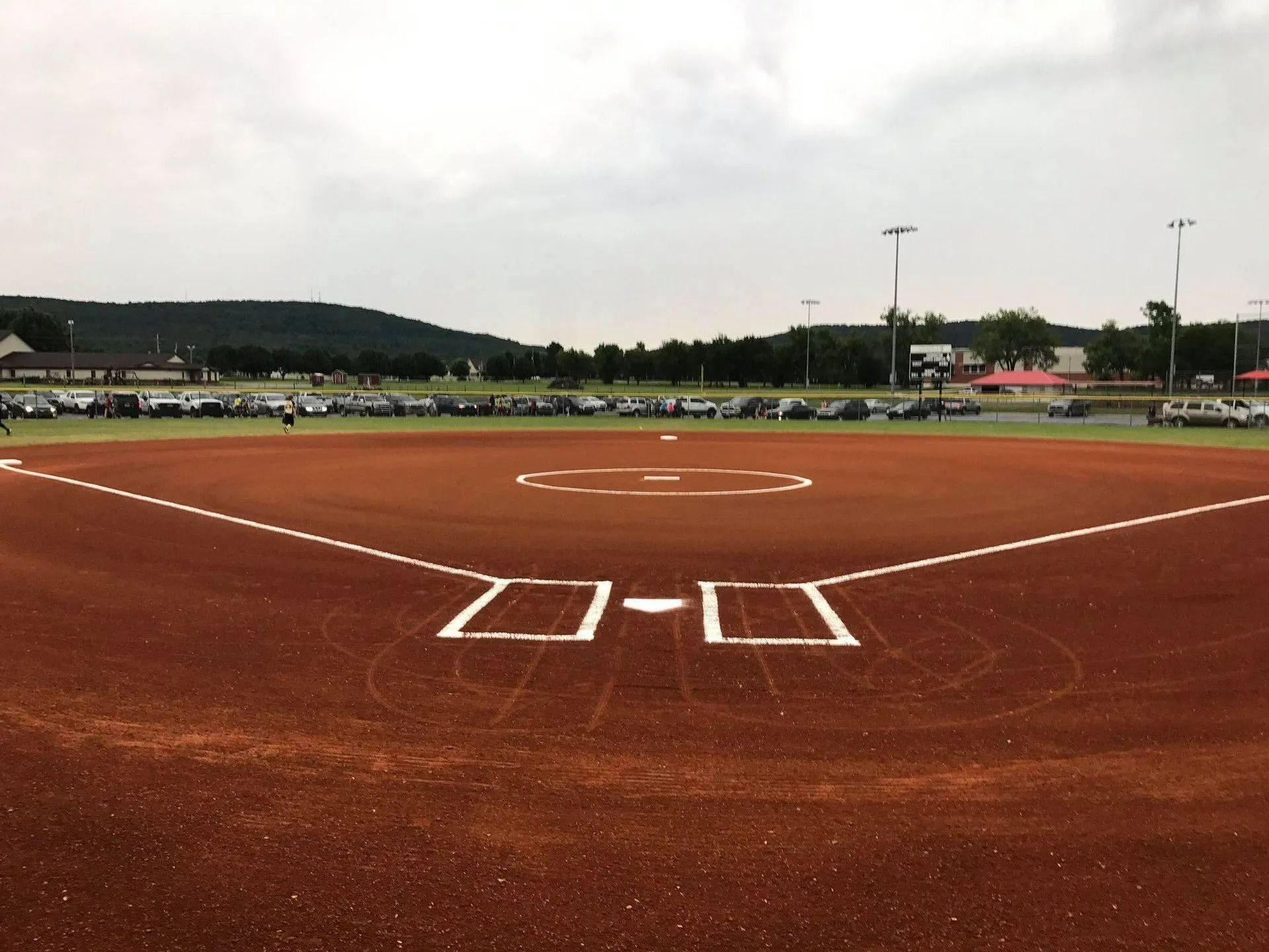 Softball field, dirt infield, white baselines, overcast sky. Cars parked beyond the outfield.