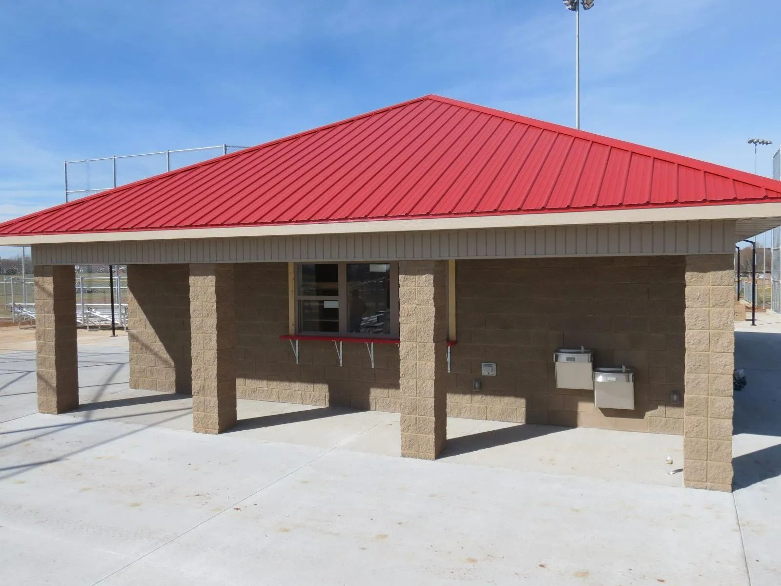 A small, open-air concession stand with a red roof. Light brown brick pillars, and a concrete floor.