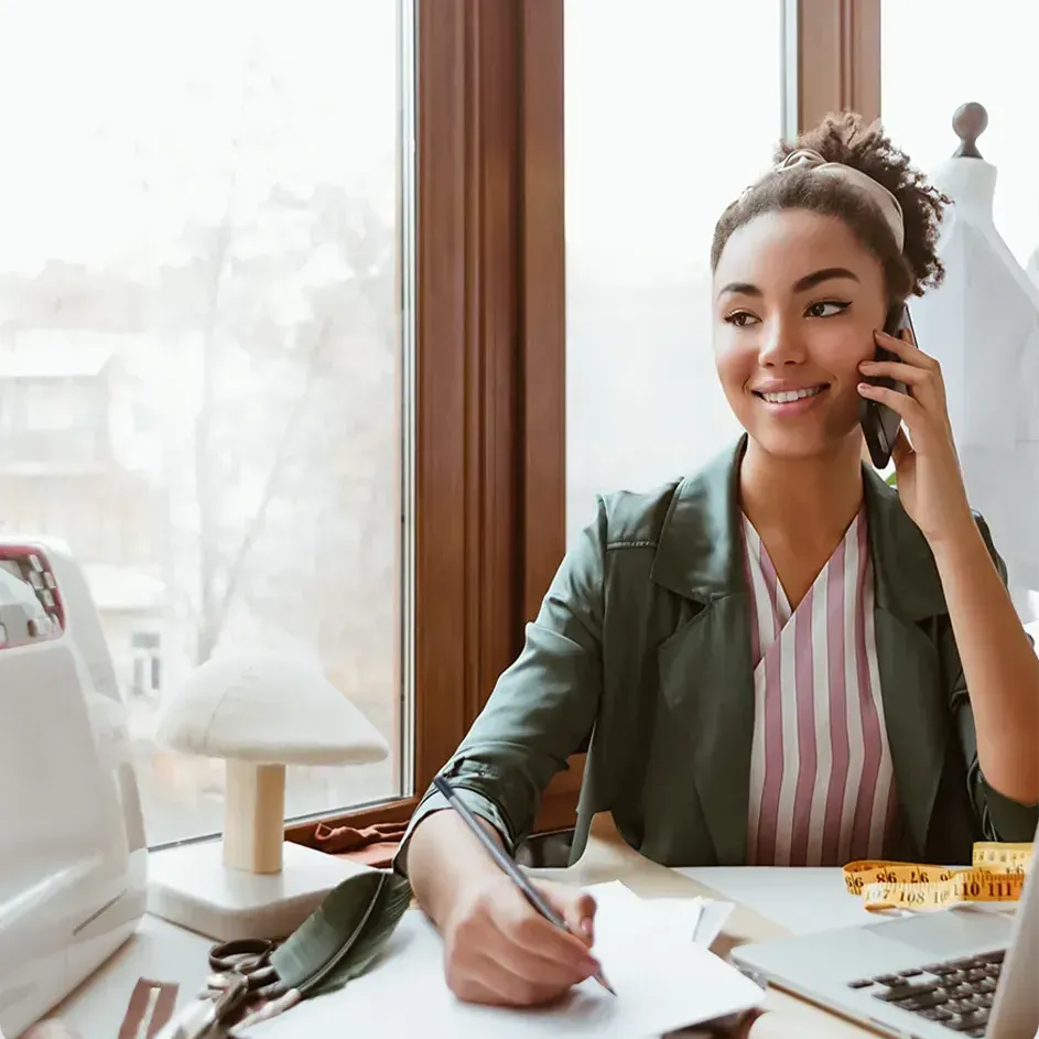 Woman in green jacket on phone, smiling, writing at desk with sewing machine and laptop.