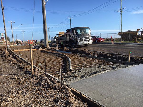 Construction site: truck pouring concrete for a sidewalk, with rebar framework visible. Sunny day.