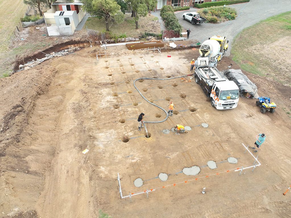 Construction site with cement being poured into drilled holes. Workers and equipment visible.