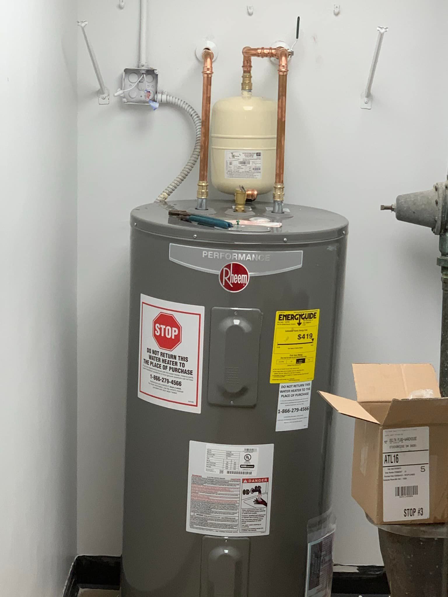 A water heater is sitting in a corner of a room next to a box.