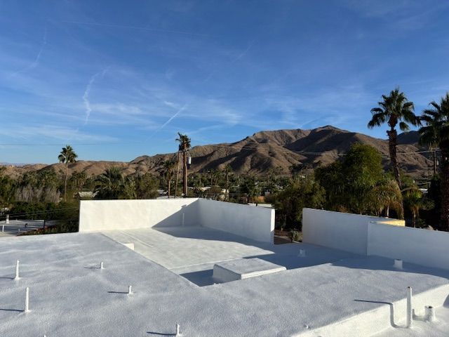 A white roof with low walls, palm trees, and mountains under a clear blue sky.