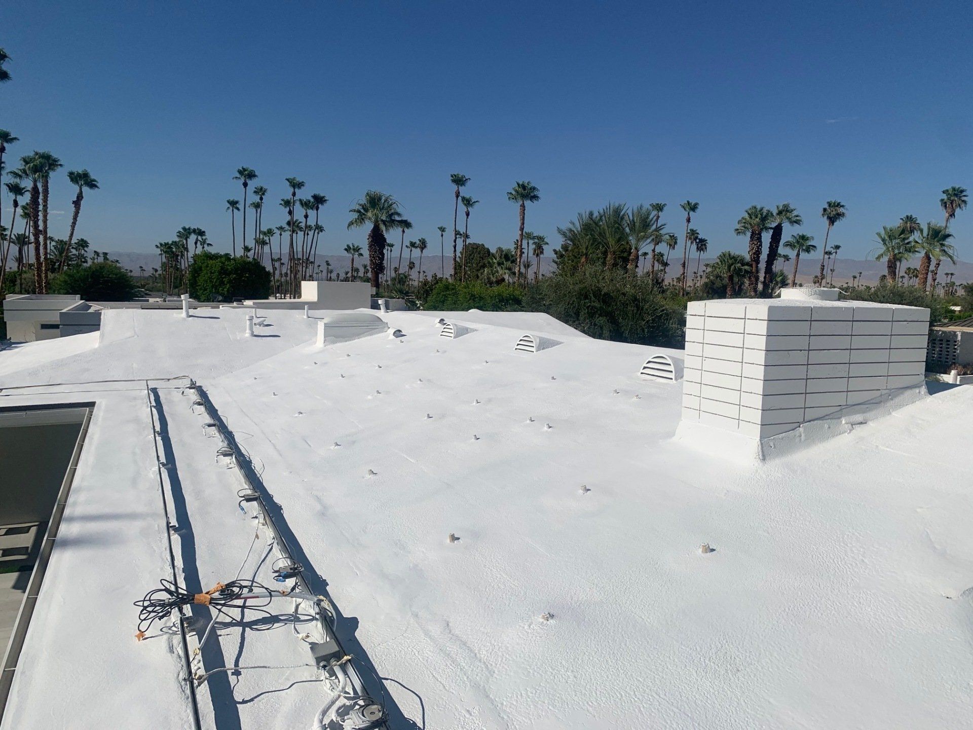 White-coated flat roof in Palm Springs, California. Chimney and solar panel tracks visible, with palm trees and mountains in the background.