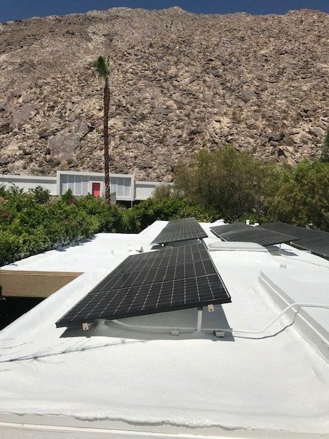 Solar panels mounted on a white roof with a mountain backdrop, a palm tree, and vegetation. Sunny outdoor setting.