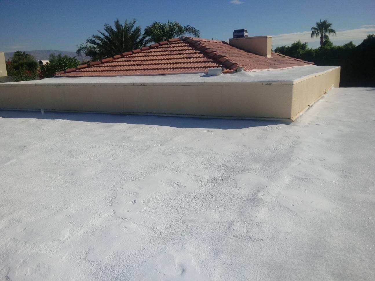 A flat roof with a light-colored coating in the foreground, a tiled roof and a building in the background under a clear sky.