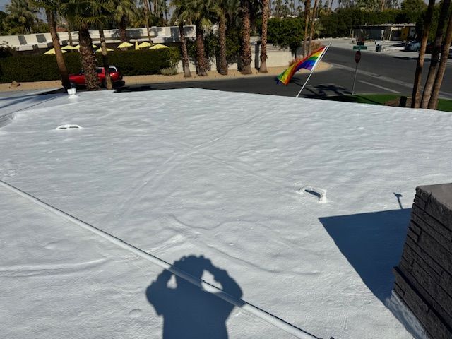 White roof with a rainbow flag waving in the background, palm trees, and the shadow of a person taking the photo.