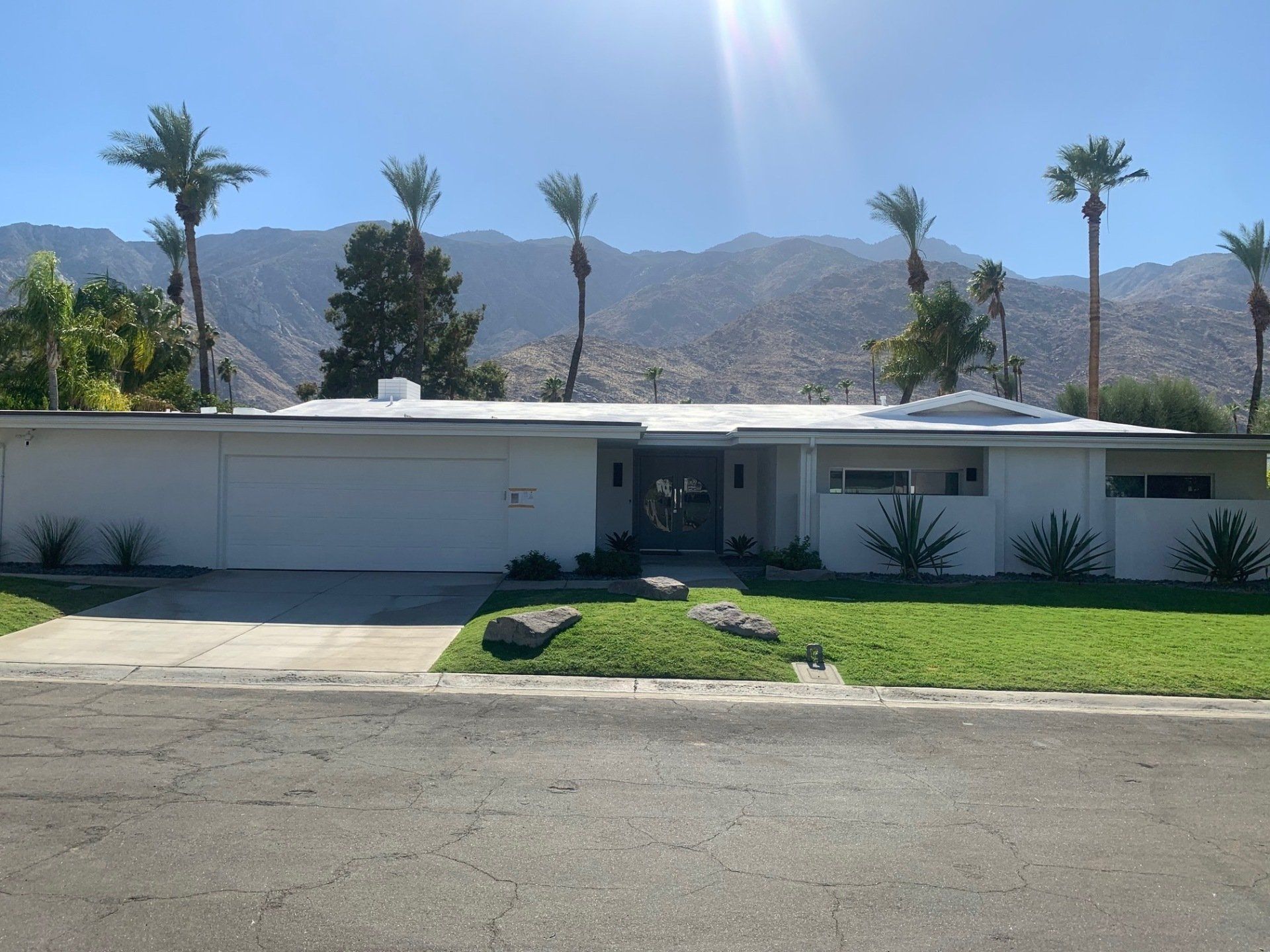 A single-story, white mid-century modern home with a flat roof, surrounded by palm trees and mountains in the background.