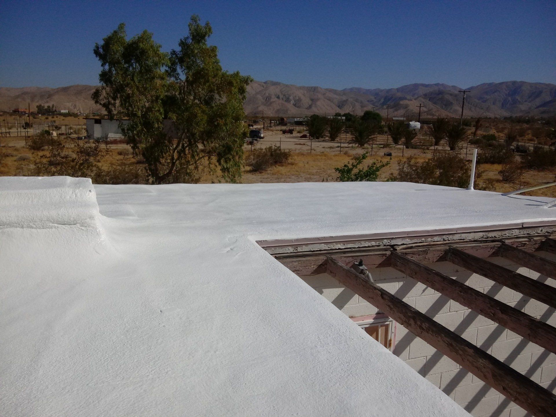 White foam roofing with a clear blue sky, mountains, and desert landscape in the background. The roof is being applied.