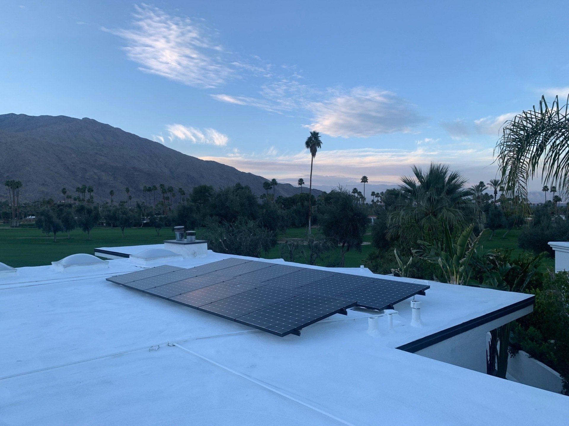 Solar panels on a white rooftop with a mountain and palm trees in the background under a blue and cloudy sky.