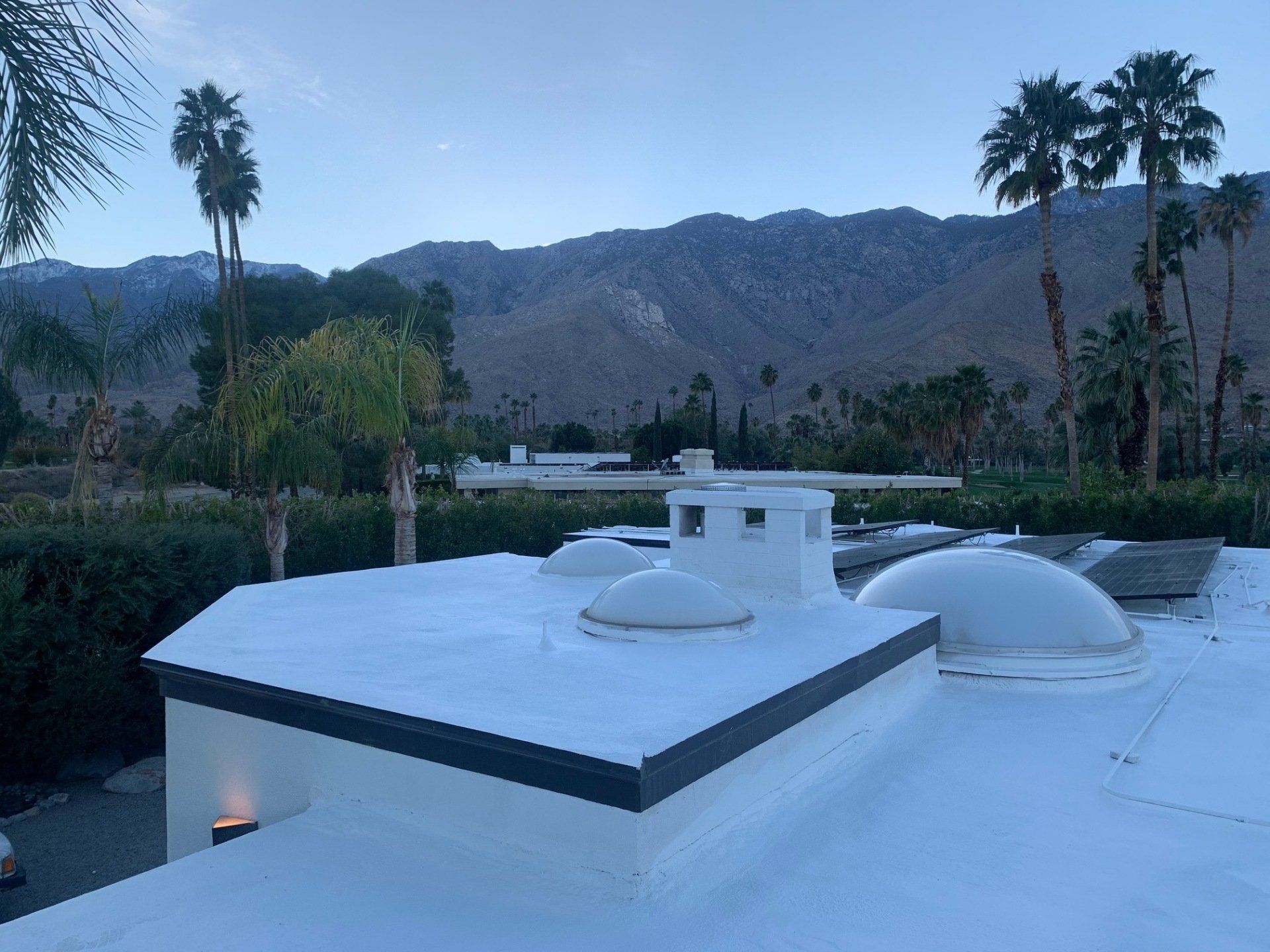 A white roof with domed skylights and a rectangular chimney faces a mountain range with palm trees in the background.