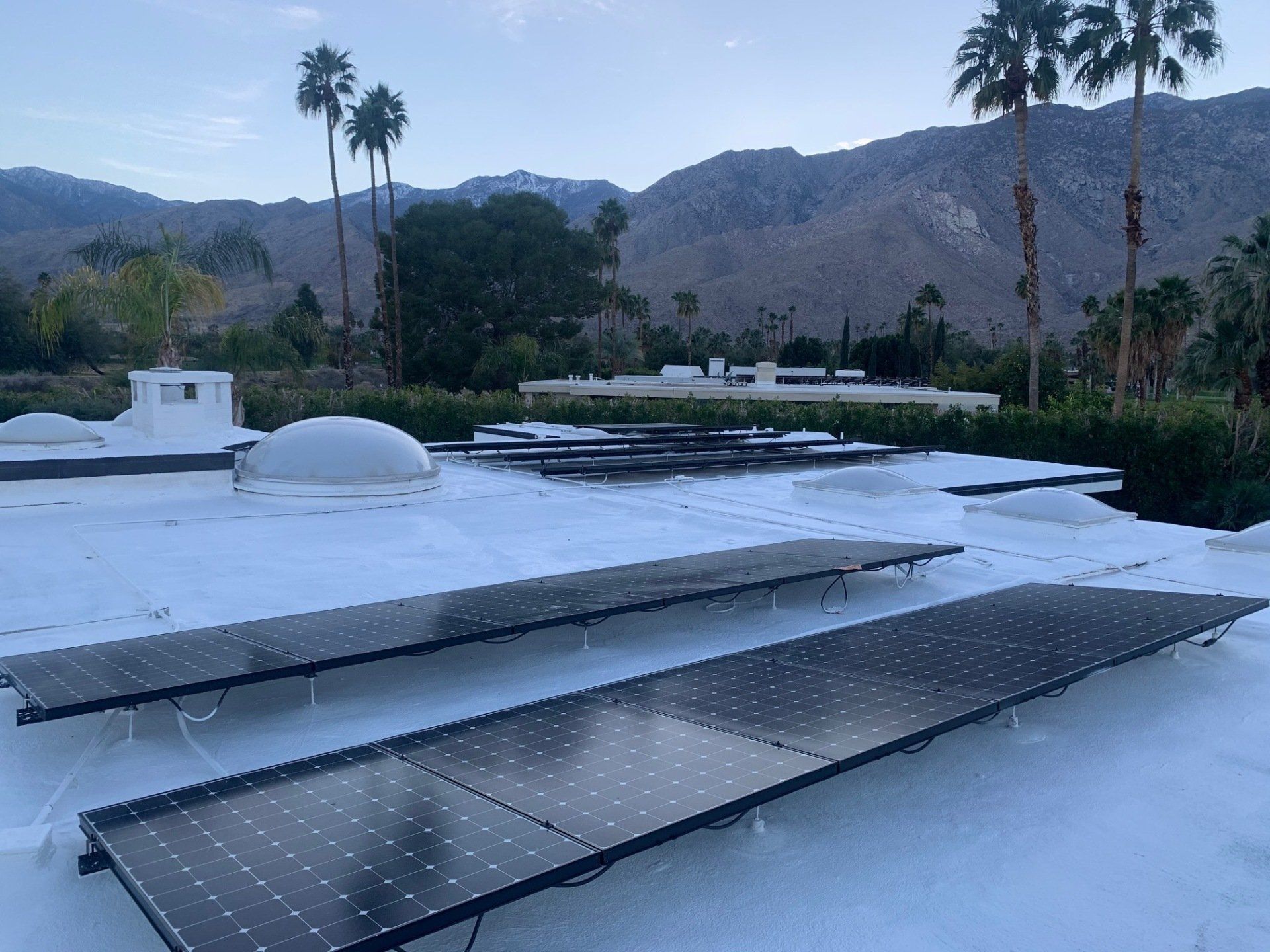Solar panels installed on a white flat roof, with mountains and palm trees in the background under a cloudy sky.
