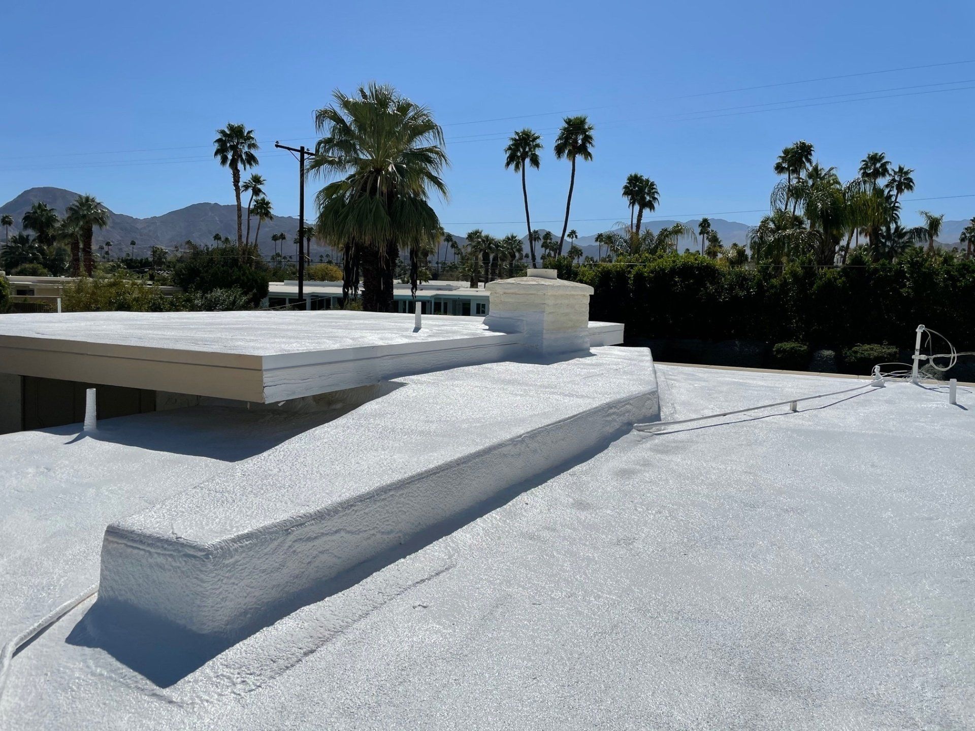 White-coated flat roof with small structure; palm trees and mountains in the background under a bright blue sky.