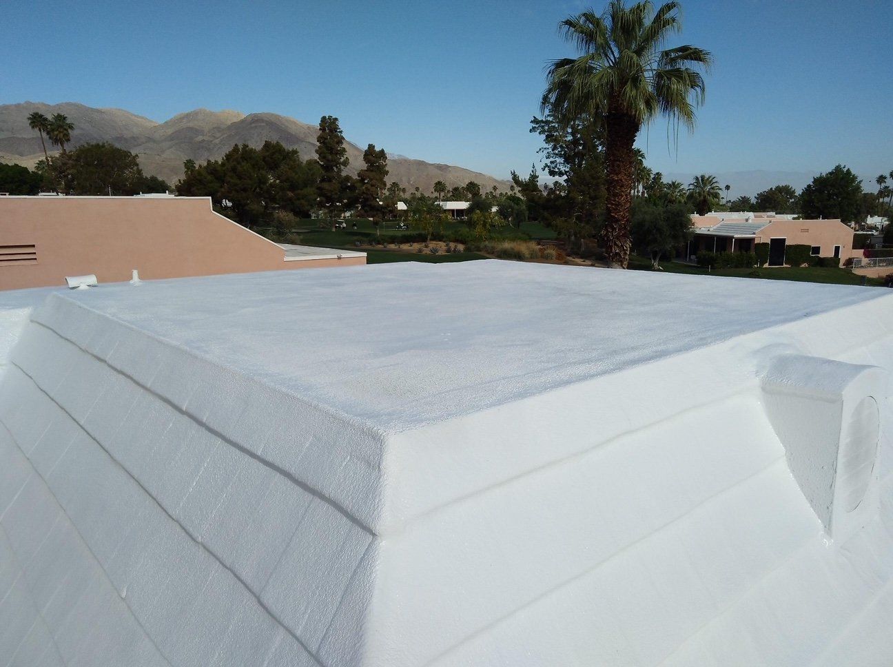 White, recently coated roof with foam insulation, in an outdoor setting with a palm tree and mountains visible in the background.