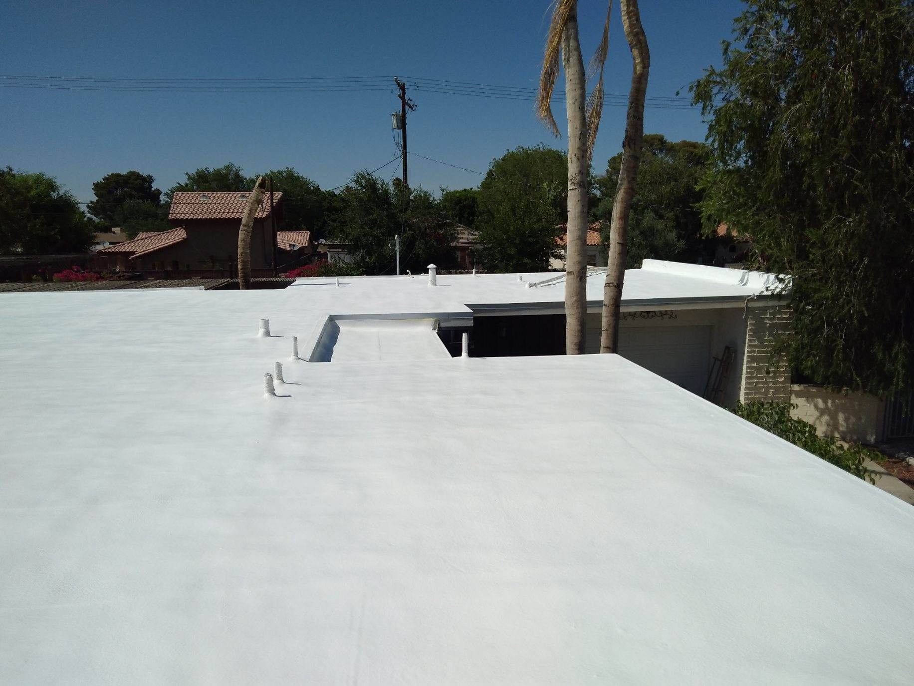 White flat roof on a building with a clear blue sky in the background. Trees and other houses are visible in the distance.