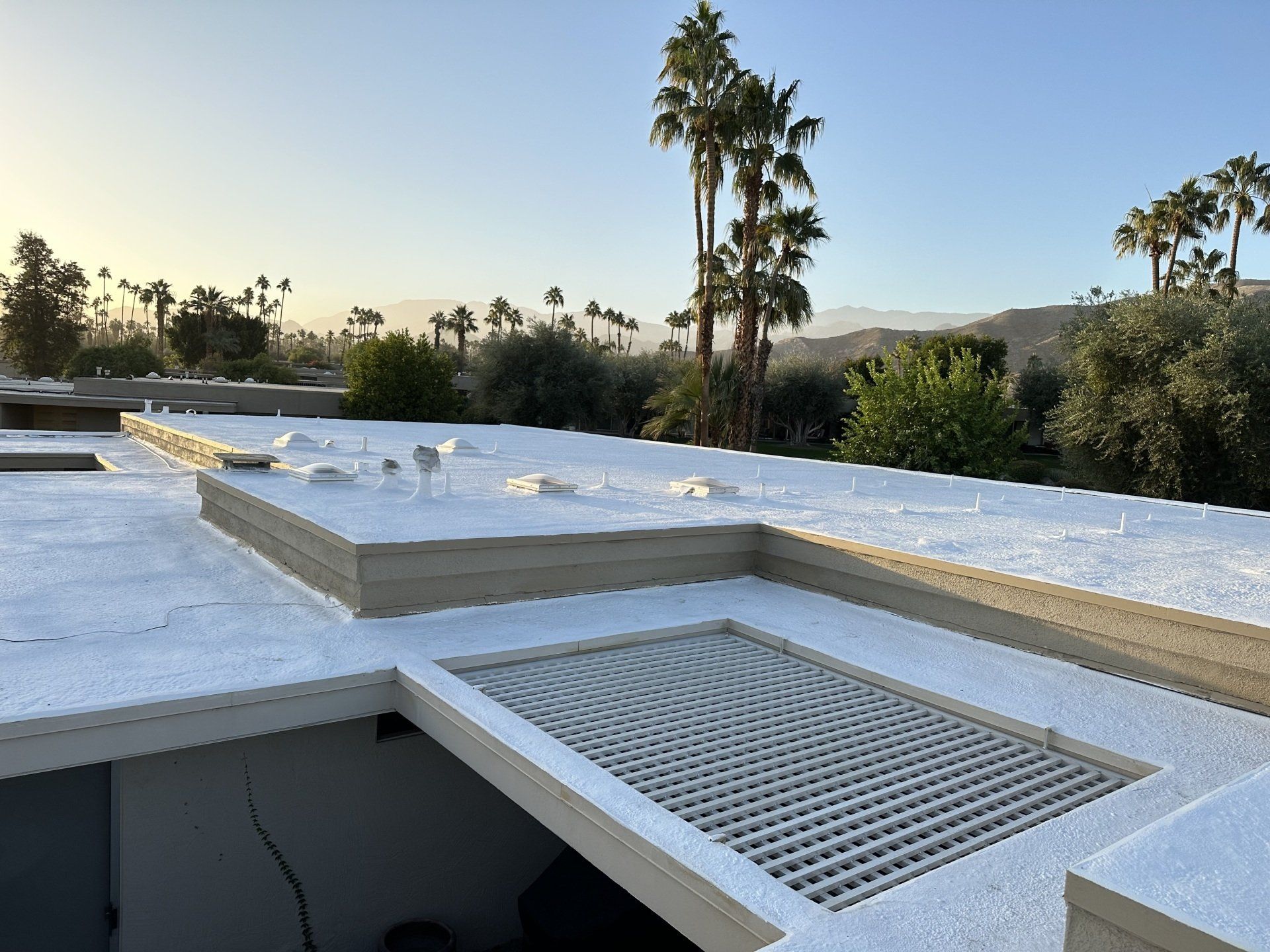 Flat, white roof with a grated skylight, surrounded by palm trees and a mountain range under a clear sky.