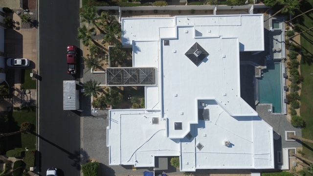 Overhead view of a modern white house with a pool, driveway, and street. Palm trees are scattered around the property.